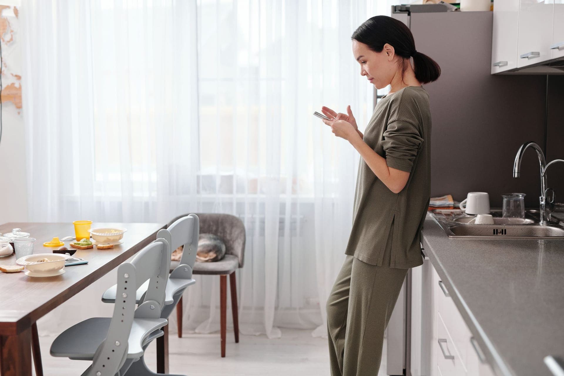 A homeowner standing in her kitchen in the morning, reading a text on her phone.