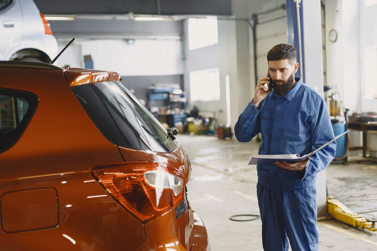 A mechanic in blue coveralls on the phone beside a customer's car in the service bay.